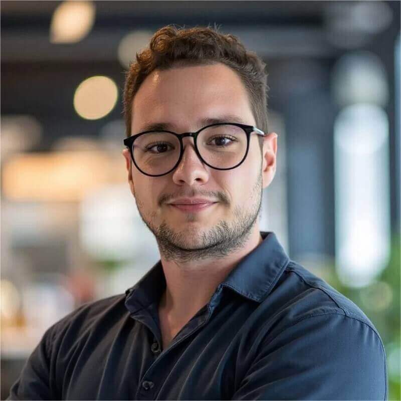 Homem branco de cabelo e barba curtos, usando óculos pretos arredondados e camisa azul escuro.
